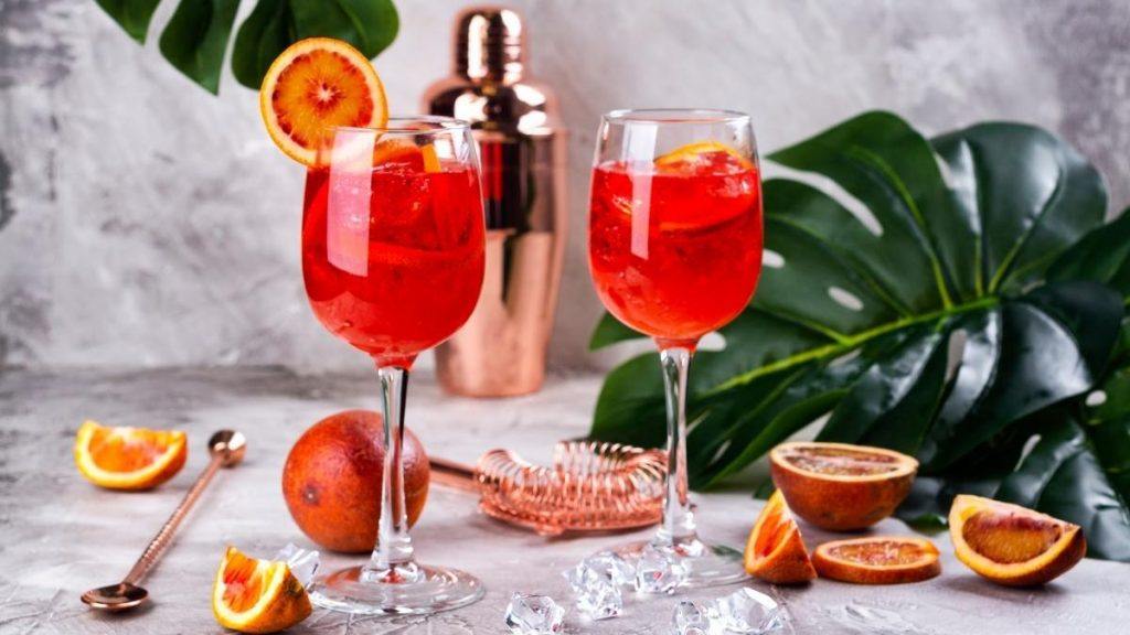 Close up of two bright red Italian Gentleman cocktails in stemmed glasses, presented against a light grey backdrop, surrounded by greenery and fresh slices of citrus fruit