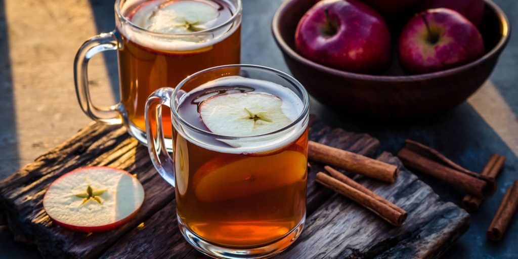 Two glass mugs of apple cider buttered rum with a bowl of apples and cinnamon sticks