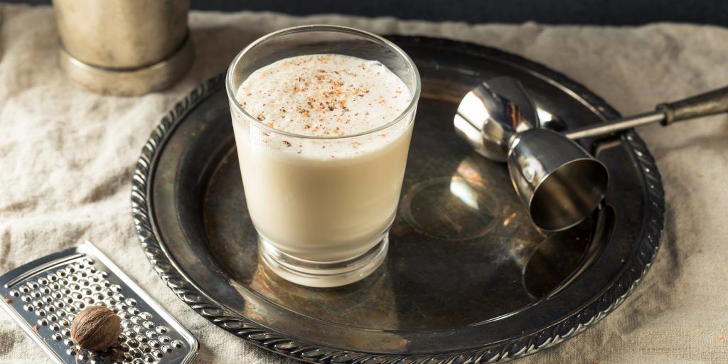Close up top shot of a creamy brandy Milk Punch cocktail in a glass tumbler, presented on a small silver platter, set on a white cotton tablecloth, with a small grater and fresh nutmeg beside it