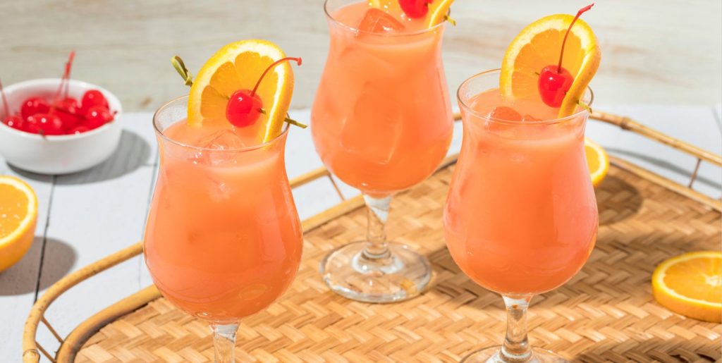 Three orange Hurricane cocktails on a bamboo tray garnished with orange slices and cherries