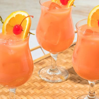 Three orange Hurricane cocktails on a bamboo tray garnished with orange slices and cherries