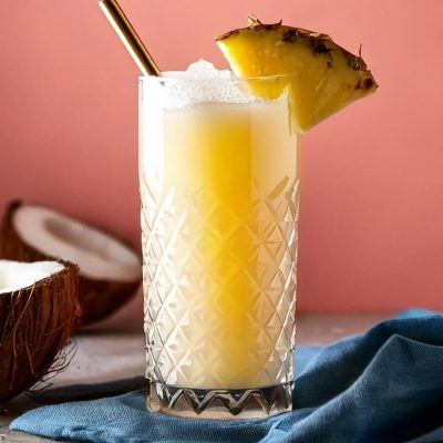 Close-up of a Painkiller cocktail with fresh pineapple wedge garnish, halved coconuts to the side, pink background