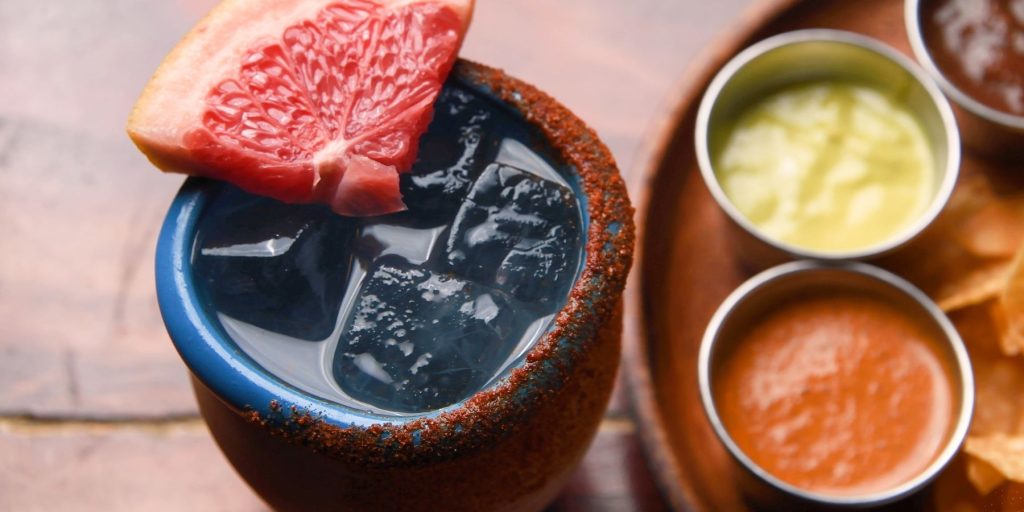 A traditional Cantarito cocktail in a terracotta cup, garnished with lime and served on a rustic wooden surface.