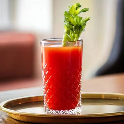 A tall glass with a deep red Bloody Mary cocktail sits on a gold tray, garnished with a fresh celery stalk. The vibrant tomato-based drink contrasts with the neutral background, adding a touch of elegance.
