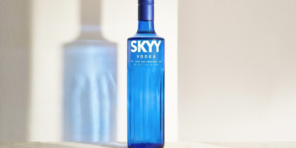 A close-up shot of a SKYY Vodka bottle with a strong shadow cast on a neutral wall, emphasizing the brand’s iconic blue glass design. The minimalist aesthetic highlights the timeless appeal of the vodka bottle. 