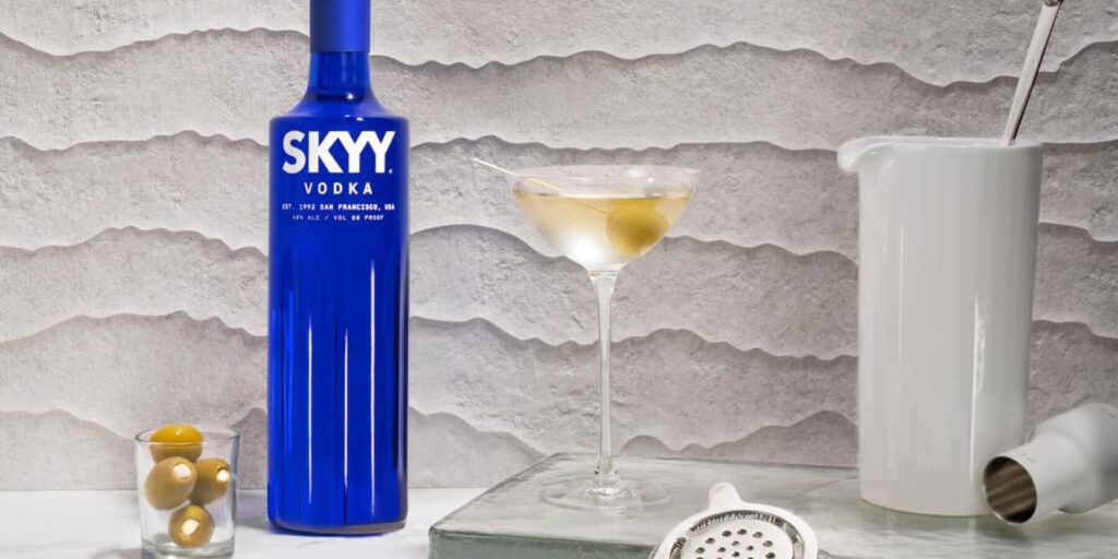 A bottle of SKYY Vodka sits beside a dirty martini garnished with a single green olive. The neutral-toned, wavy-textured backdrop and neatly placed bar tools emphasize a refined cocktail aesthetic.