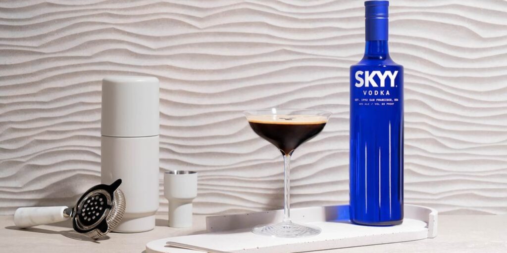 A bottle of SKYY Vodka is displayed alongside an espresso martini, with a foamy top and rich, dark color. The modern, textured backdrop and sleek bar accessories add a contemporary, stylish touch.