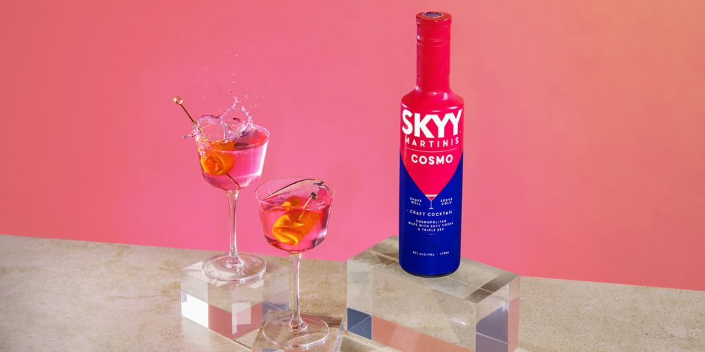 A stylish SKYY Cosmo Martini bottle with two Cosmopolitan cocktails splashing in glasses.