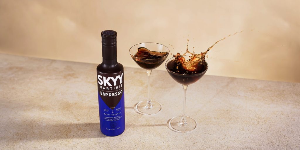 A SKYY Espresso Martini bottle with two Espresso Martinis, one mid-splash, against a beige background.