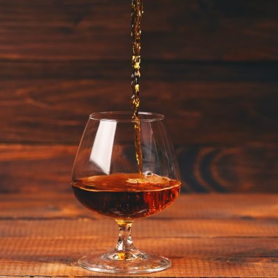 Pouring shot of cognac into a glass on a wooden table, photo by levgenii_Meyer