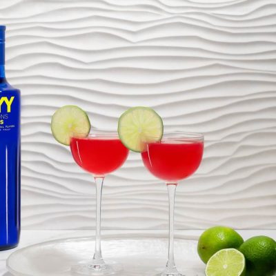 A vibrant cosmopolitan cocktail with lime garnishes sits next to a SKYY Vodka bottle, set against a white textured background. The red cocktail pops against the clean, modern setting, embodying a chic, feminine vibe.