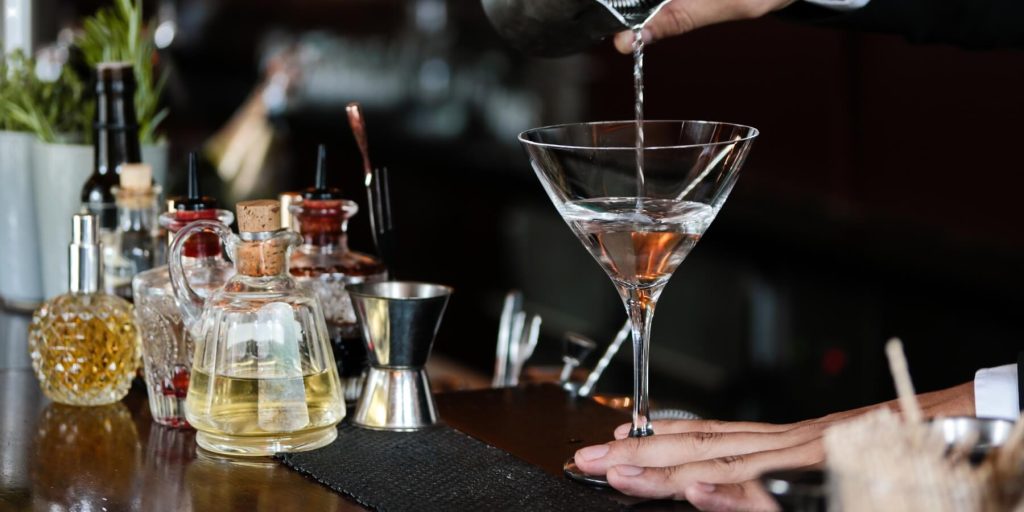 A bartender pours a Perfect Martini into a V-shaped cocktail glass at a professional bar setup, surrounded by bitters, syrups, and bar tools.