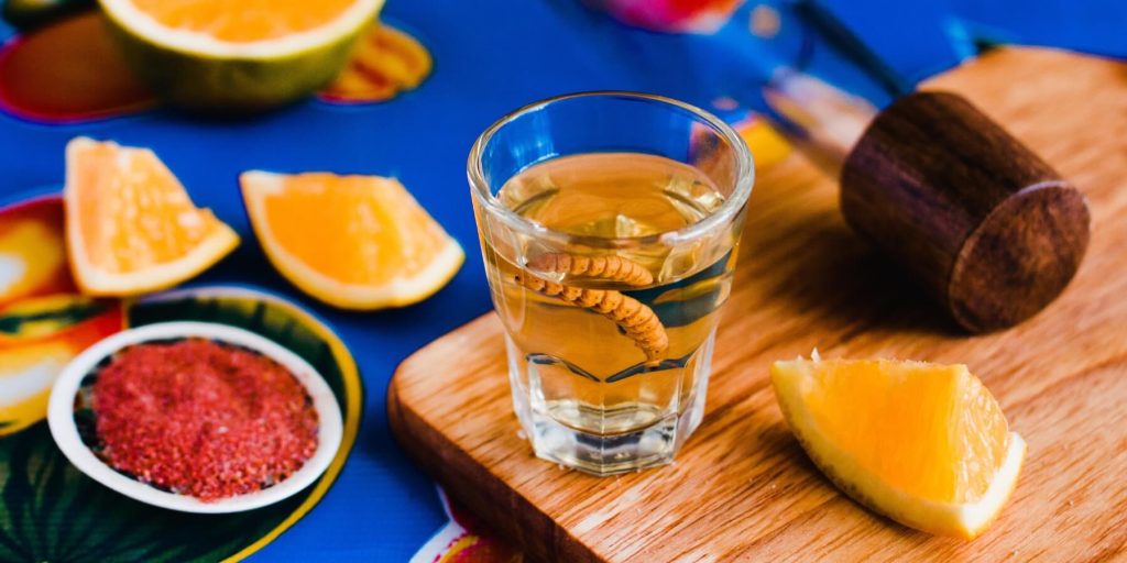 Close-up of a gusano in a shot glass of mezcal, surrounded by orange slices and a salt rim, photo by eskymaks/Adobe Stock