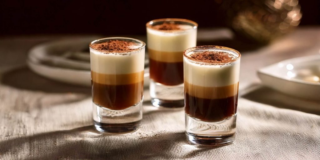 Three layered Tiramisu Shots in shot glasses, featuring dark coffee, creamy liqueur, and a dusting of cocoa powder on top.