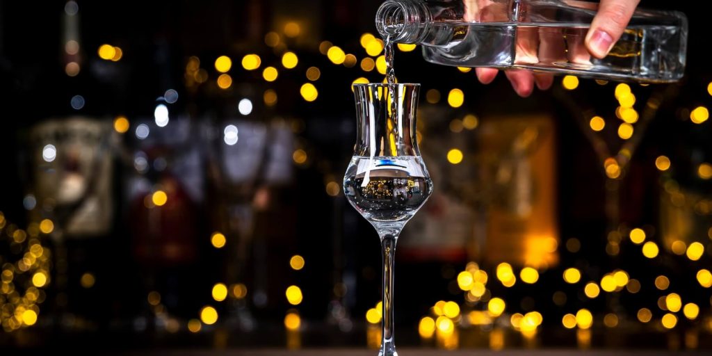 Clear grappa being poured into a traditional narrow glass with golden bokeh lights in the background, photo by 5ph/Adobe Stoc