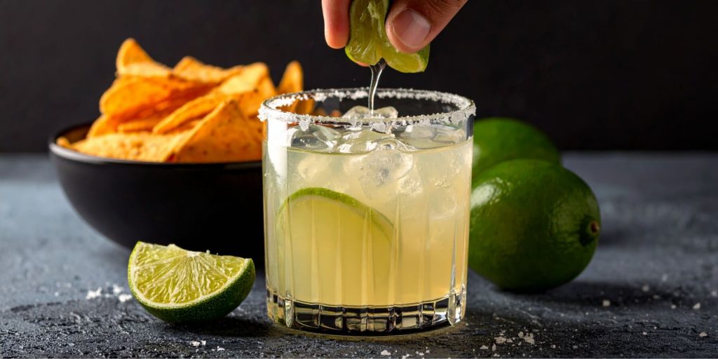 Close-up of a hand squeezing a halved lime with the juice dripping into a Margarita cocktail