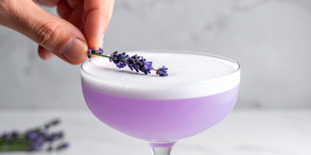 Close-up of a hand placing a lavender sprig on a foamy Lavender Gin Fizz cocktail in a coupe glass.