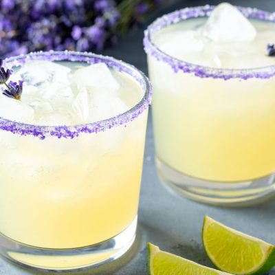 Two Lavender Margaritas with purple sugar rims and lavender sprigs, surrounded by lime wedges and fresh lavender.