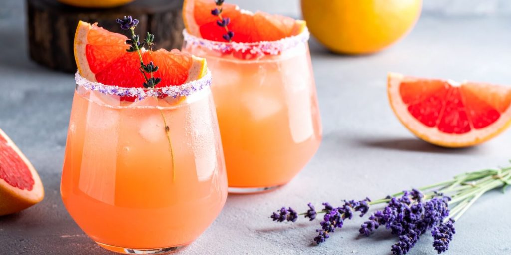 Two Lavender Paloma cocktails garnished with grapefruit wedges and lavender, with sugared rims and citrus slices in the background.