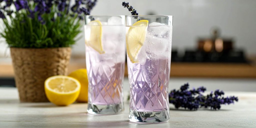 Two tall glasses of Lavender Vodka Cooler with lemon slices and ice, set beside fresh lavender and lemon halves.