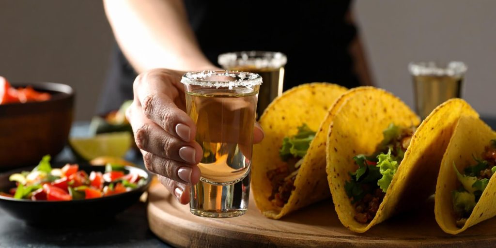 Close-up of a woman's hand holding a glass of tequila, tacos to the side