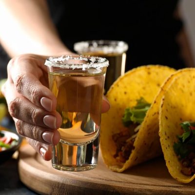 Close-up of a woman's hand holding a glass of tequila, tacos to the side