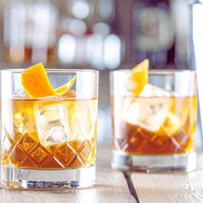 Two whiskey Old Fashioned cocktails on a bar counter with bottles of bourbon in the background