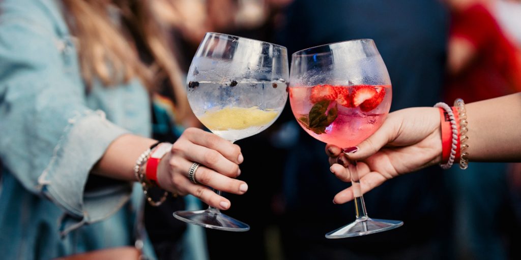 Two people clinking balloon glasses filled with vibrant gin and tonic variations, one with lemon and juniper berries, the other with strawberries and mint, at an outdoor event; image by @kiomu.studio via Adobe Stock.