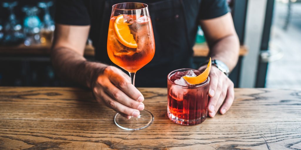 Bartender in a black T-shirt holding a short glass of Campari and a tall glass of Aperol