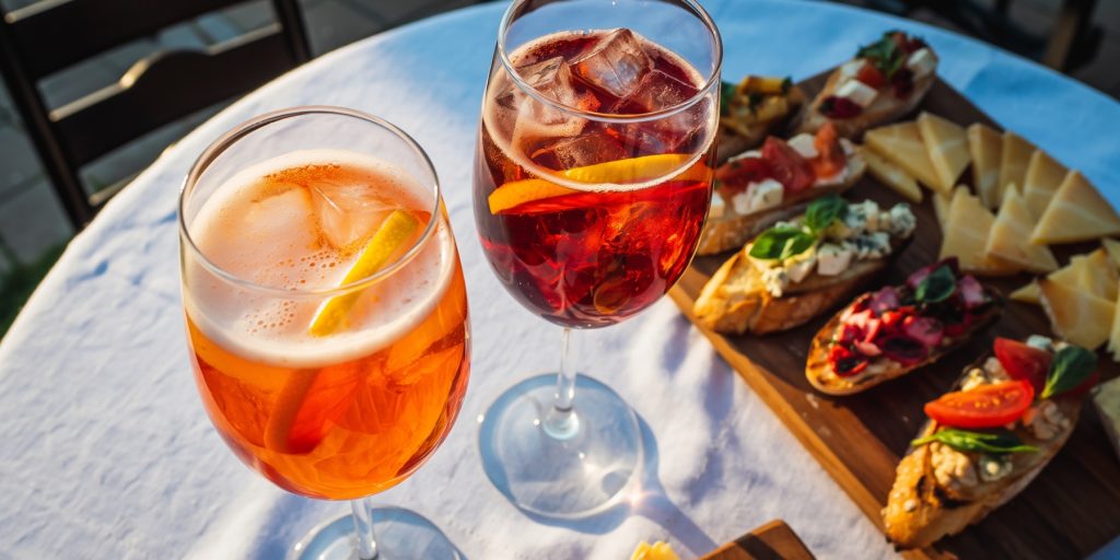 A glass of Aperol on the left and a glass of Campari on the right set on a table with a platter of bruschetta and cheese