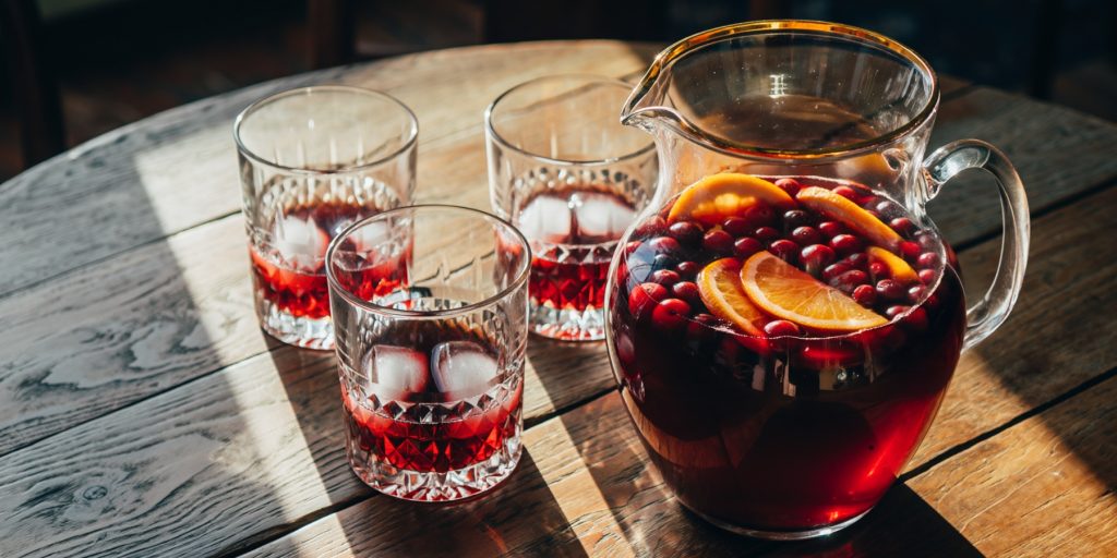 A glass pitcher of Thanksgiving Sangria, a deep red batch cocktail brimming with cranberries and orange slices. Three matching glasses with ice are arranged nearby on a wooden table in dappled sunlight.