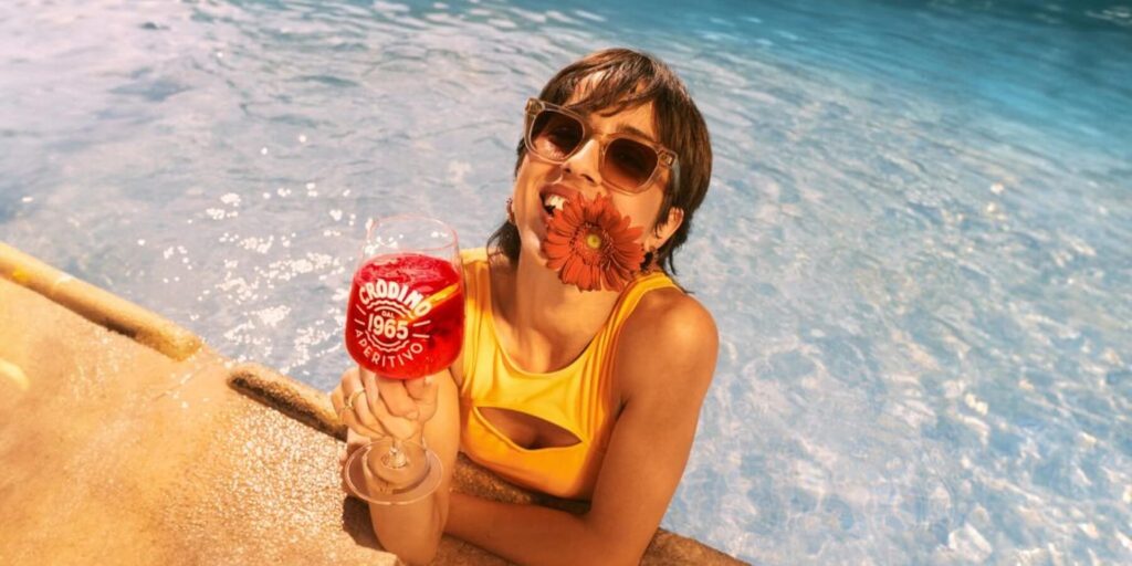 A young lady with sunglasses on and a flower in her teeth drinking a glass of non-alcoholic Rosso Crodino