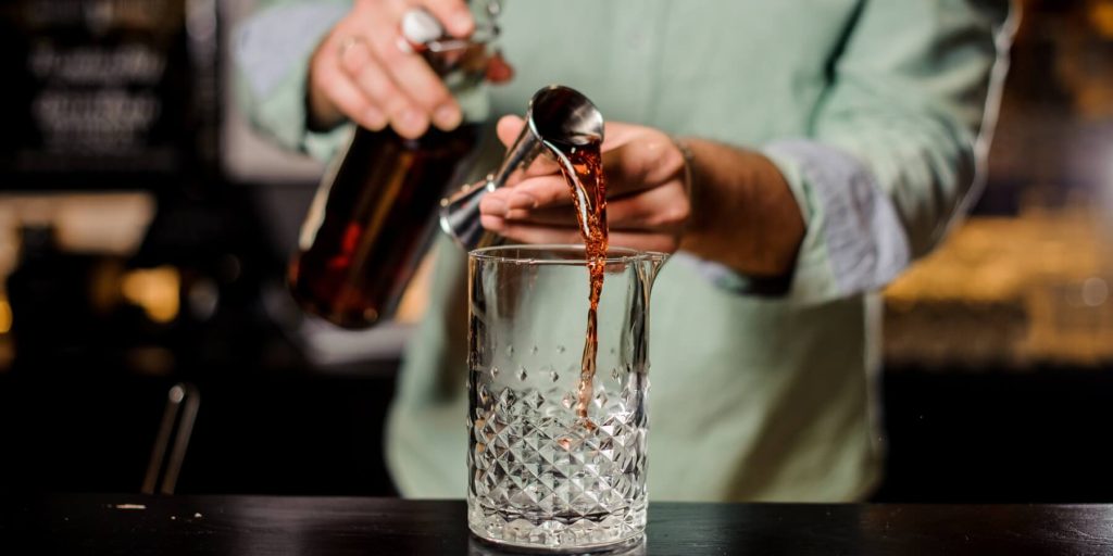A bartender pouring a measure of spirit out jigger into a glass mixing jug