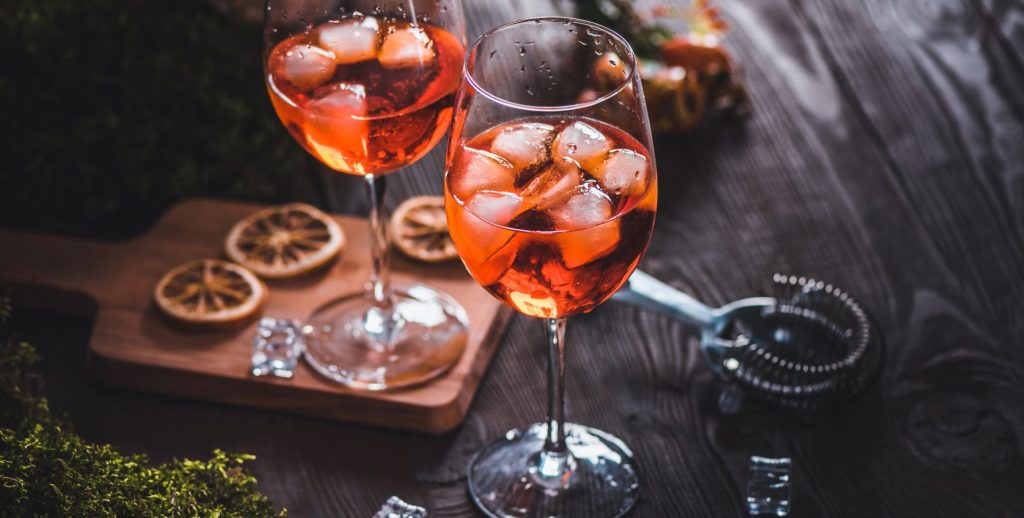 Two Aperol spritz cocktails on a wooden surface surrounded by fresh thyme and dried orange wheels.