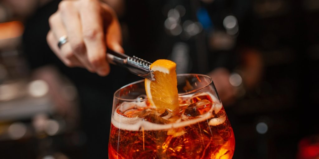 A close up shot of a bartender garnishing an Aperol spritz cocktail with a fresh orange slice using tweezers.