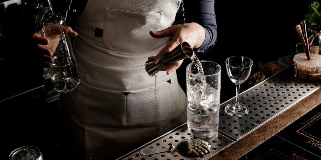 Close up image of a female bartender pouring a shot of gin into a Collins glass over ice in a commercial bar, lots of natural light
