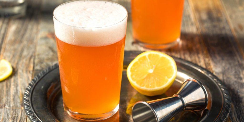 Close up of two Aperol Mist beer cocktails presented on a small silver serving tray alongside a metal jigger and half a lemon