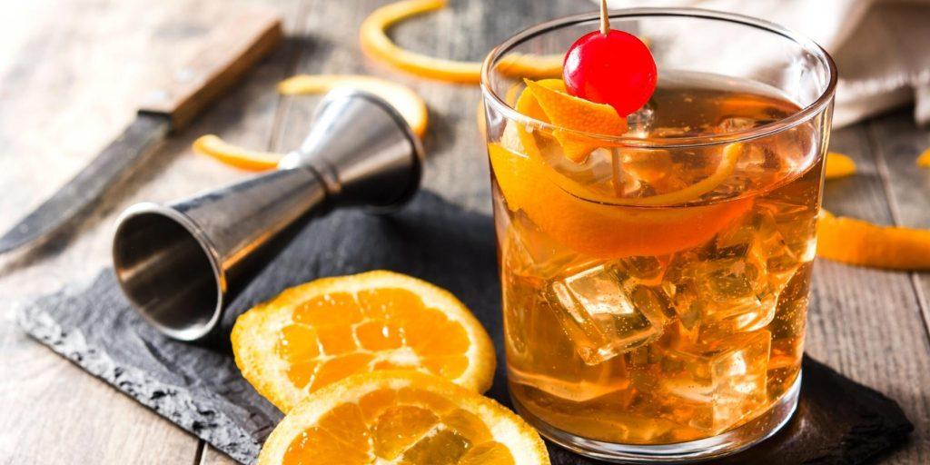 Aperol Old Fashioned with orange slice cherry garnish presented on a rustic wooden surface alongside some orange slices and a metal jigger