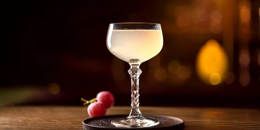 A crystal coupe glass filled with the Indian Summer cocktail, placed on a wooden surface with a few fresh red grapes nearby and a warm, moody background.