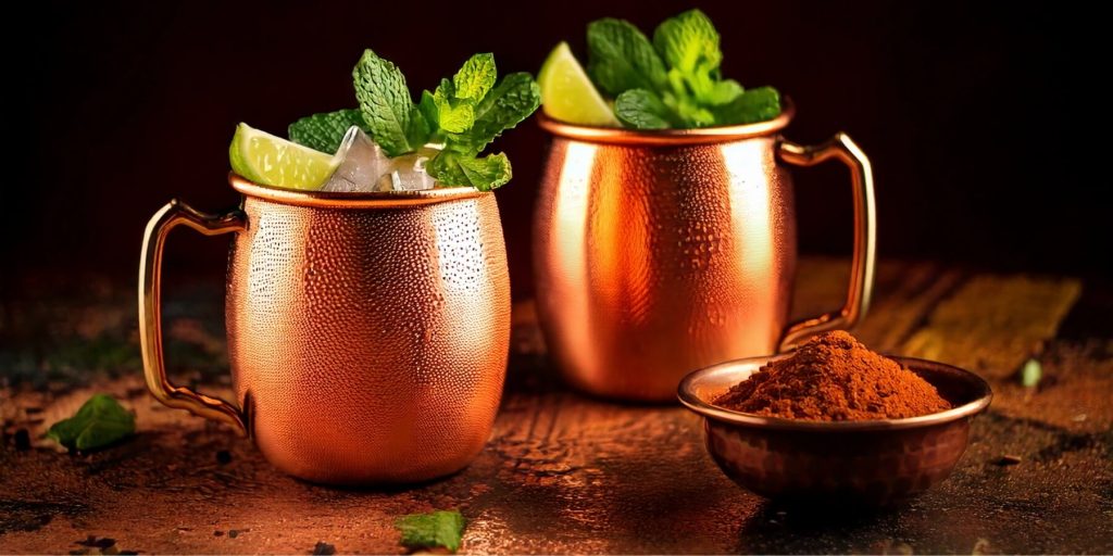 Two hammered copper mugs filled with the Mumbai Mule cocktail, garnished with mint sprigs and lime wedges, next to a small copper bowl of powdered spices.
