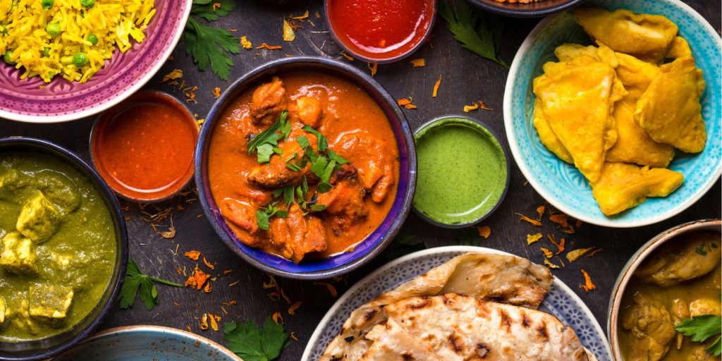 A colourful Indian food spread featuring butter chicken, palak paneer, yellow rice with peas, pakoras, naan, and a variety of vibrant chutneys and sauces, all served in patterned bowls and plates.