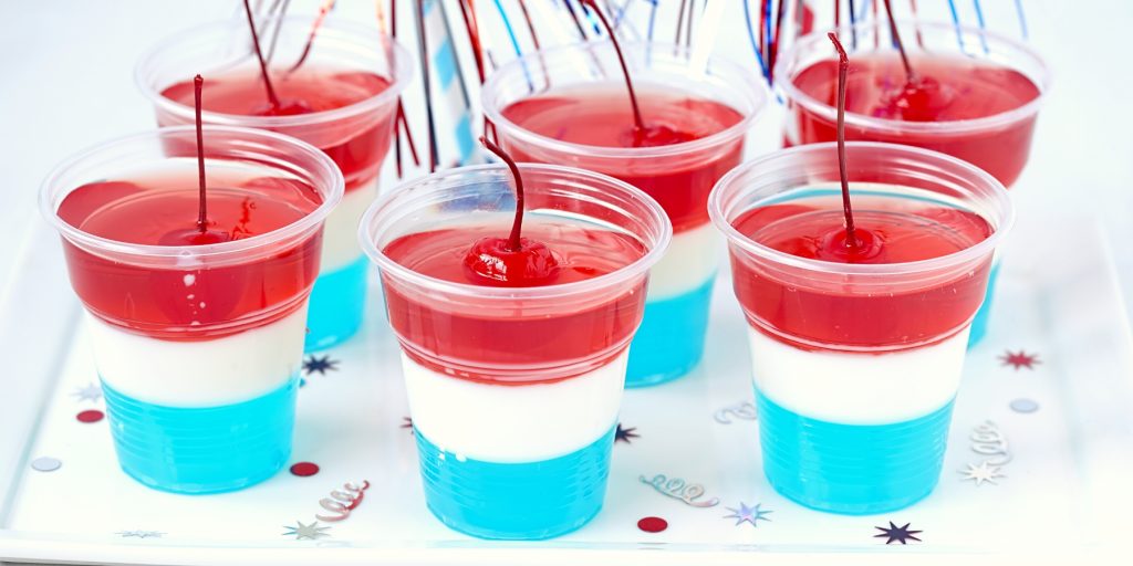 Close up of a collection of blue, white and red layered jello shots in plastic shot glasses, garnished with red cherries