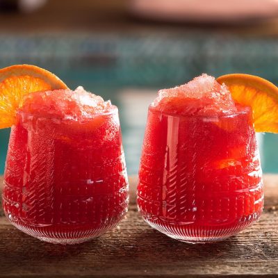 Close-up of two bright red Frozen Negroni cocktails with orange garnish, served poolside