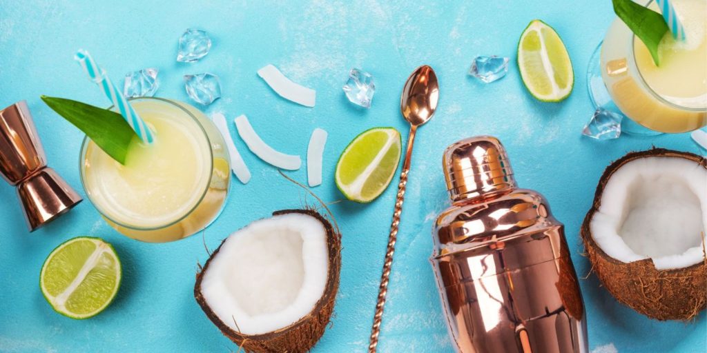 Top view of all the ingredients and tools required to make a Pina Colada cocktail, on a light blue backdrop in a flat lay format