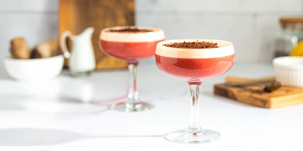 A rich, dark cherry-red Black Forest Gateau cocktail served in a coupe glass, topped with a creamy foam layer and sprinkled with chocolate shavings. 