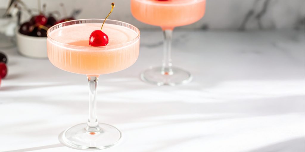 A pale pink Cherry Alexander cocktail served in a coupe glass, garnished with a single red cherry.