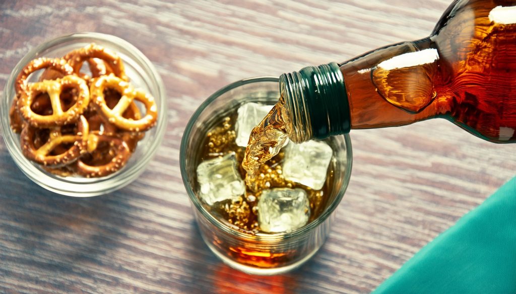 A top view of bourbon being poured into an ice filled glass, bowl of pretzels next to it
