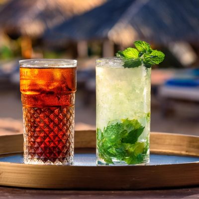 A Cuba Libre and a Mojito on a tray in a beach setting