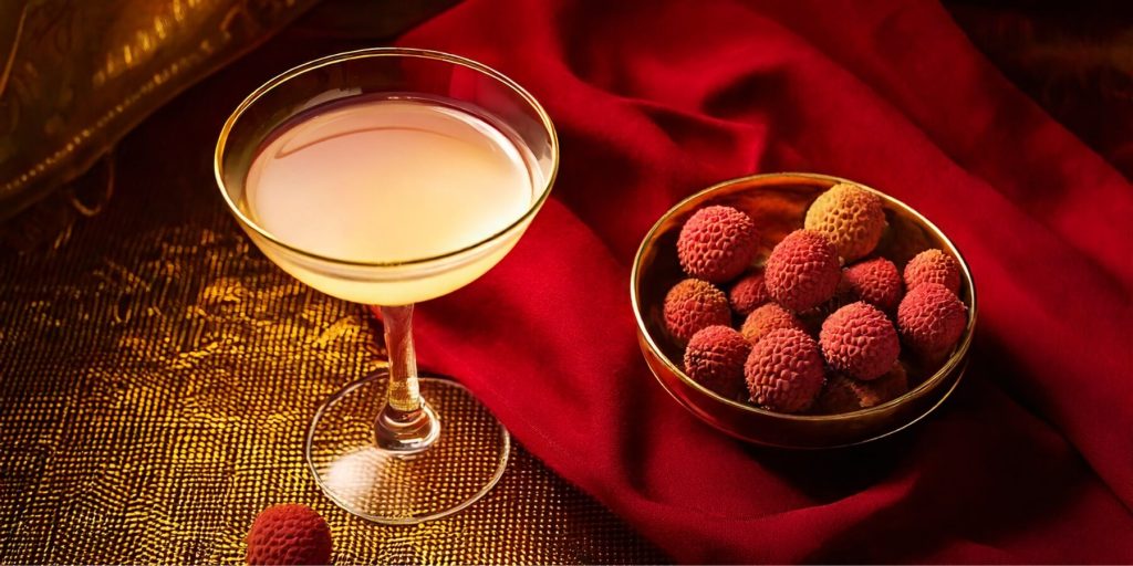 A delicate cocktail in a coupe glass accompanied by a bowl of fresh lychees, set against a rich red fabric and golden accents.