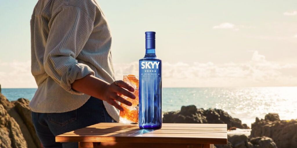A person enjoys a cocktail on a beachside setting with a bottle of SKYY Vodka placed on a wooden table. The golden sunlight and ocean waves in the background create a relaxed atmosphere.